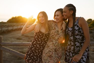 Positive multiracial female best friends smiling brightly and hugging each other while leaning on fence on wooden pier at sundown
